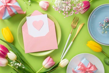Moda Anneler Günü kutlaması. Renkli üst manzara tabaklar, açık zarf içinde kartpostal, çatal bıçak, hediye, vazo ve laleler açık yeşil arka planda.