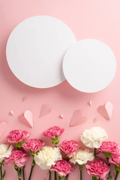 Mother\'s Day concept. Top vertical view flat lay of elegant carnation flowers, and pink paper hearts on a soft pastel pink background with two blank hearts for advert