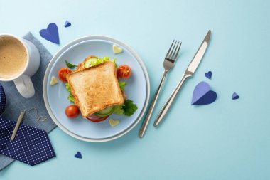 Babamı özel bir Babalar Günü kahvaltısıyla şımart. Sebzeli, bıçaklı, çatallı, kahve kupalı, peçeteli, kravat iğneli, erkek aksesuarlı, pastel mavi arka planlı ev yapımı bir sandviç.