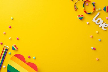 Gökkuşağı broşları, bileklik ve kalp şeklindeki kart da dahil olmak üzere gurur temalı aksesuarların bulunduğu düz bir daire, canlı sarı bir arkaplanda, LGBT Tarih Ayı temasını kutluyoruz.