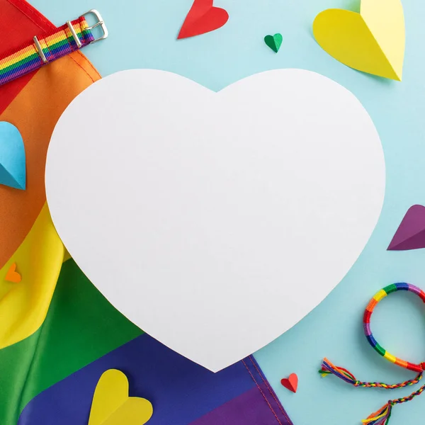 stock image Vertical square top view of LGBT pride accessories, including a rainbow flag, wristlet, pin badges, on a pastel blue background with an empty heart for ad, representing the LGBT History Month concept