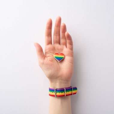 Sembolik gökkuşağı renkli bileziğin birinci şahıs kare görüntüsü, LGBTQ Tarihi Ayı onuruna beyaz arka planda kalp şeklinde bir rozet taşıyan bir kadını süslüyor.