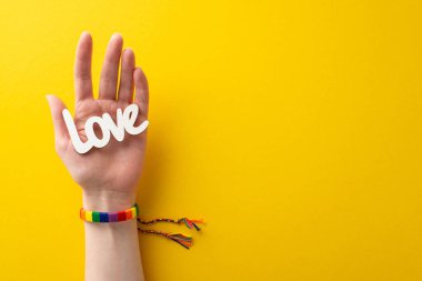 Gökkuşağı renginde bir bilezik takmış bir kızın elini ilk gören kişi, açık sarı bir arkaplanda, mesaj ya da reklam için boş bir arkaplanda, LGBTQ Tarih Ayı 'nı tanıyan bir yazı tutar.