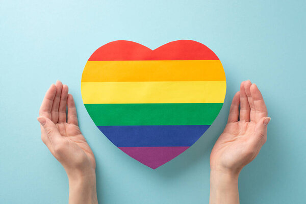 Embodying the spirit of LGBT History Month, this image features woman's hands showing a rainbow heart-shaped card on pastel blue background with empty space for text or advertising