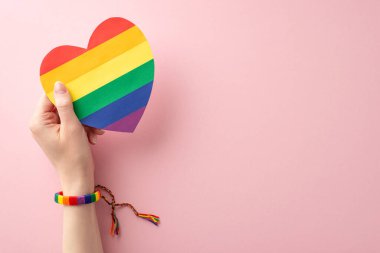 LGBT Tarih Ayı teması: Sembolik bilezik içinde genç bir kadının el fotoğrafı, Pastel pembe arka planda gökkuşağı kalp kartı, metin veya reklam için boşluk.