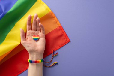 Sembolik gökkuşağı renkli bileziğin birinci şahıs görüntüsü, LGBTQ Tarih Ayı 'nı tanıyan, Leylak arkaplan üzerinde kalp şeklinde bir rozet taşıyan bir kızın elini süslüyor.