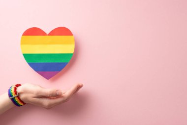 Kadın elinin sembolik bir bilezik taktığı ve pastel pembe arka planda gökkuşağı kalbi çıkardığı fotoğraf. LGBT geçit töreni konsepti için mükemmel.