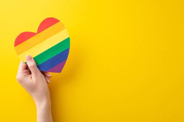 LGBT Tarih Ayı teması: Metin veya reklam için boş alanı olan canlı sarı arkaplan üzerinde gökkuşağı kalp kartpostalı tutan genç kadının ilk kişi görüntüsü