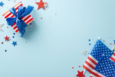4 Temmuz kutlama teması. Parlayan yıldızlar, canlı konfeti, Amerikan bayrakları, hediye kutusu, mavi zemine karşı dizilmiş, metin veya reklam için boş alan da dahil olmak üzere parti dekorasyonlarının en üst görüntüsü