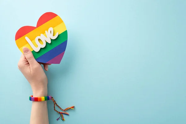 stock image First person top view photo of young woman's hand wearing symbolic bracelet holding LGBTQ+ flag card with text love on pastel blue background with space for ad copy