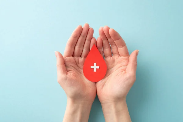 A first-person perspective top view photo of a female hands holding blood droplet with cross sign on palms on pastel blue background. It\'s the perfect visual for raising awareness about blood donation