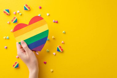 LGBT Tarih Ayı fikri: genç bir kadının gökkuşağı kalp kartını sarı arkaplandaki geçit aksesuarlarının üzerinde tutarken çekilmiş ilk kişi fotoğrafı