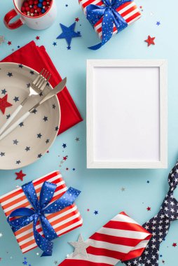 Büyüleyici bir masa düzenlemesiyle 4 Temmuz 'u yükseltin! Masa takımlarının, çatal bıçakların, konfetilerin ve açık mavi yüzeyin dikey üst görüntüsü. Boş fotoğraf çerçevesini metin veya resimle özelleştir