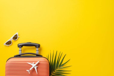 Rüya gezisi konsepti. Altında palmiye yaprağı olan turuncu bavulun üst görüntüsü ve güneş gözlüğü yanında fotokopi alanı olan izole edilmiş parlak sarı arka planda uçak modeli.