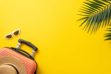 Yaz tatili konsepti. Üstünde hasır şapka ve palmiye yaprakları olan turuncu bavul ve güneş gözlüklerinin fotosu izole edilmiş, sarı arka planda, kopyalanmış.