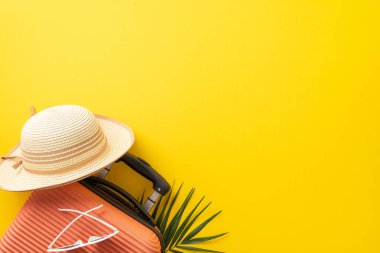 Yaz tatili kavramı. Palmiye yaprağının altında turuncu bir valizin ve üzerinde güneş gözlüğü olan hasır bir şapkanın, kopyalanmış sarı arka planda çekilmiş yüksek görünüm fotoğrafı.