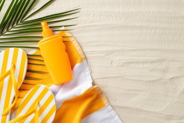 Tatil kavramında güneş koruması. Plaj havlusunun en üstteki fotoğrafı, sarı parmak arası terlikler, güneş kremi spreyi ve kopyalanmış arka planda kumun üzerindeki palmiye yaprağı.