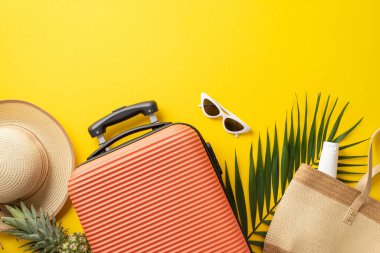Tropik tatil konsepti. Hasır şapka ve çantayla çevrili turuncu bavulun en üstteki fotoğrafı, izole edilmiş, parlak sarı arka planda, fotokopi alanı olan palmiye yaprağı ve güneş gözlüğü.