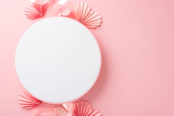 Top view photo of empty white circle with a pastel pink ribbon and paper origami hearts on an isolated light pink background with copyspace