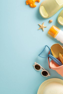 Çocuk konseptiyle tatil. Çocuk plaj oyuncakları, panama, parmak arası terlik ve güneş gözlüğü, güneş kremi spreyi ve mavi izole arka planda kabukları olan boş bir alanın dikey fotoğrafı.