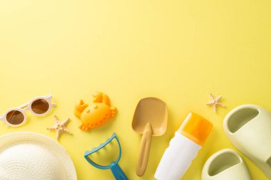 Çocuk konseptiyle tatil. Yukarıda, kum, güneş kremi, terlik, güneş gözlüğü, panama şapkası ve sarı izole arka planda deniz kabukları için çocuk kumsalı oyuncaklarıyla dolu boş bir yer fotoğrafı var.