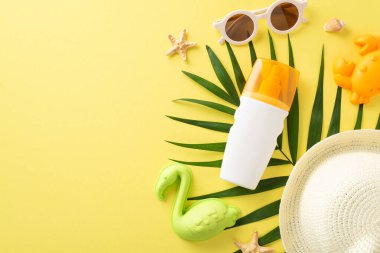 Çocuklu tropik bir tatil köyü. Palmiye yaprağı ve kabukları, çocuk kum oyuncakları, güneş kremi şişesi, güneş gözlüğü ve sarı izole arkaplan üzerine panama içeren boş bir alanın en üstteki resmi.