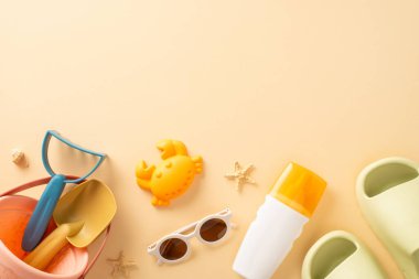 Bebekle plaj kaçamağı: oyuncakların yukarıdan aşağı görünümü, kumdan kale aletleri, güneş kremi şişesi, gözlük, lastik terlik, kabuk, sakinleştirici bej arka planda deniz yıldızı, mesaj veya reklam için yer sunma