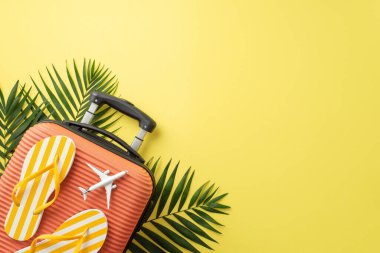 Tatil titreşimleri kaydedildi. Turuncu bir valizin üst görüntüsü, plaj aksesuarları, parmak arası terlikler, ve yeşil palmiye yaprakları sarı zemin üzerinde mesaj veya promosyon için boş alan.