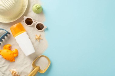 Çocuk konsepti için plaj aktiviteleri. Yüksek açılı görüntü, güneş şapkası, güneş gözlüğü ve güneş kremi şişesi ile kumdaki kumları ve deniz kabuklarını yakalayarak izole edilmiş mavi arka planda kopyalanmış alan sunuyor.
