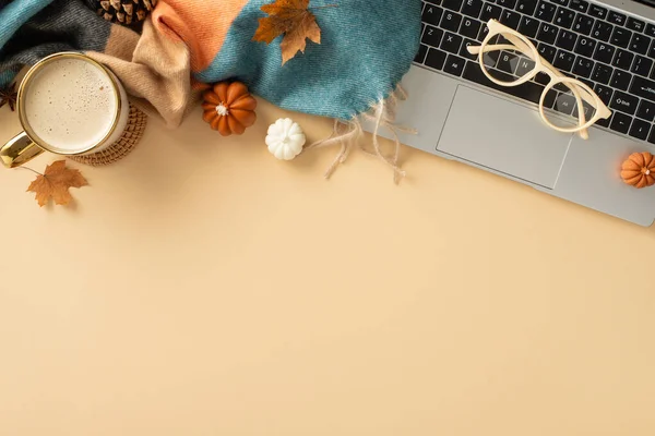 Autumn-themed desk setup. Top view of laptop, steaming mug of fresh coffee, reading glasses, cozy blanket, miniature pumpkins, golden leaves, pine cone, pastel beige backdrop with space for text or ad