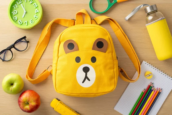 Primary education concept. Top-view image of assorted supplies, notepad, color pencils, sharpener, glasses, clock, toy, magnifier, playful bear-shaped backpack, water bottle, and apples on wooden desk