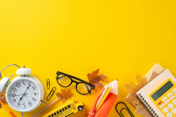 Organize your study space: top view various school supplies like copybooks, pencil case, calculator, clock and more. Vibrant yellow background, decorated with maple leaves, perfect for promo message