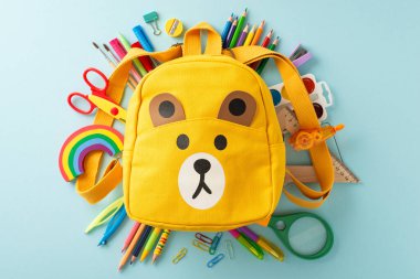 Küçük çocuklar için eğitim. Sarı çocuk sırt çantasının üst görüntüsü. Kalemler, kalemler, suluboya boyalar, makas ve mavi izole edilmiş arka plan gibi çeşitli renkli okul malzemeleriyle dolu.