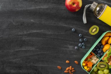 Okul için dengeli atıştırma fikri. Yukarıdan aşağıya görünen plastik beslenme çantası. Taze meyve, sandviç, böğürtlen, fındık, su şişesi, karatahta zemin üzerine yerleştirilmiş. Mesaj veya reklam içeriği için boşluk.