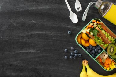 Dengeli okul atıştırmalığı için fikir. Beslenme çantasının üst görüntüsü sağlıklı seçenekler içeriyor. Meyve, sandviç, çatal bıçak ve daha fazlası da dahil. Tahta zemine karşı sunulan, metin veya reklam için ideal