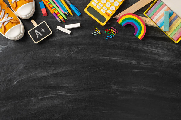 Tools for artistic exploration. Top view of colored pencils, pens, chalk set, A+ reward sticker, ruler, clips, calculator, plasticine, sporty sneakers on a chalkboard background for custom text