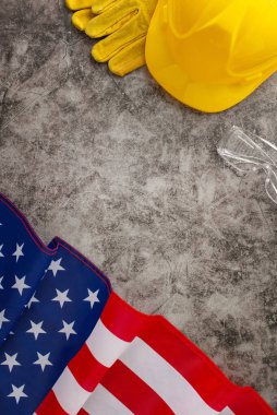 İşçi Bayramı 'nda inşaat işçilerinin sıkı çalışmalarını onurlandırmak. Amerikan bayrağını gösteren tepeden tırnağa görüntü, sarı kask ve grunge desenli gri beton üzerinde çalışma eldivenleri. Reklam veya metin için uygun
