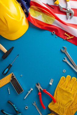 İşçi Bayramı marangozluk konsepti. ABD bayrağının dikey üst görüntüsü, çekiç, çiviler, vidalar, pense, boru anahtarı, fındık, iş eldivenleri, güvenlik gözlükleri, miğfer, mavi zemin, marangozluk dersleri için yer