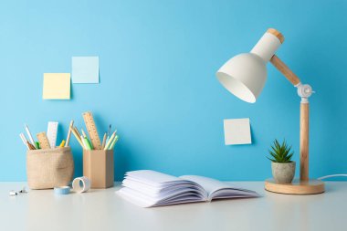 Okul için masa başı malzemelerinin yan görüntüsü: kalem organizatörleri, kalem cetvelleri, açık defter, lamba, çiçekli saksı, yapışkan notlar ve bant. Mavi duvar arkaplanı, metin veya reklam entegrasyonu için mükemmel