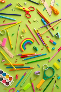 Okula dönüş temasını vurgulayın: Dağılmış renkli kalemlerin dikey üst görünümü, suluboya boyalar, plasticin, makas, cetvel, klipler, büyüteç, silgi ve pastel yeşil arka plan üzerine daha fazlası
