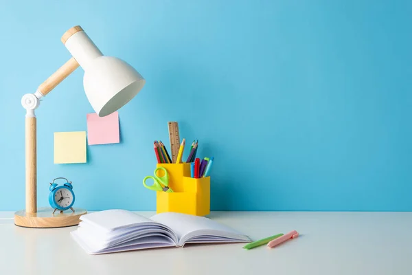 School time inspiration: side view photo of a desk adorned with school supplies, stationery holder, open book, and alarm clock. Blue wall backdrop perfect for text or ads