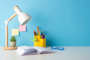 Sınıf ayarları: Okul malzemelerinin düzgün yerleştirilmiş yan görüş fotoğrafı. Kalemler, cetvel, açık kitap, yapışkan notlar, lamba, bardaklar aranjmana eklenir. Mavi duvar arkaplanı metin ya da reklam eklemeye izin verir