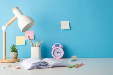 Okul zamanı konsepti yan görünüm fotoğrafına kaydedildi, masa başları gösteriliyor: kalem tutacağı, cetvel, kitap, çalar saat, lamba, saksı, yapışkan notlar, bant. Metin veya reklam fırsatı içeren mavi duvar arkaplanı