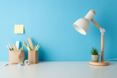 Okul masasının temel eşyalarının yan görünümü: kalem organizatörleri, cetvel, lamba, saksı, yapışkan not ve yapıştırıcı bant, güzel bir şekilde mavi duvara yapıştırılmış, metin veya reklam için alan