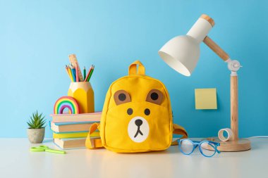 Sınıf teması. Düzgün düzenlenmiş masa başı okul malzemelerinin yan görüntüsü: kırtasiye standı, kitaplar, sırt çantası, bardaklar, lamba, saksı ve daha fazlası. Mavi duvar arkaplanı, metin veya reklamlar için mükemmel