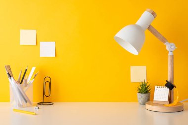 Okulun yaratıcılık düzenlemesi. Kalem standı, cetvel, klip şeklindeki kitap tutucu, takvim, küçük akademik şapka, bitki, lamba, yapışkan notlar, sarı duvar arkaplanı. Metin veya reklam için boşluk