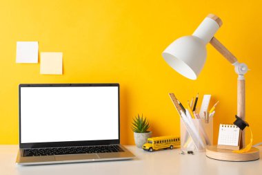 Organize çalışma alanı. Kalem tutacağı, kalem tutacağı, cetvel, takvim, küçük akademik şapka, bitki, dizüstü bilgisayar, lamba, okul servisi oyuncağı, sarı duvara yapıştırılmış yapışkan notlar, tanıtım için yer