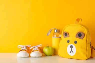 Küçük çocuklar için neşeli bir okul zamanı. Turuncu sırt çantası, spor ayakkabısı, su şişesi ve elmanın sarı izole zemin üzerindeki yan görüntüsü. Metin yerleştirme veya reklam için fotokopi alanı var.