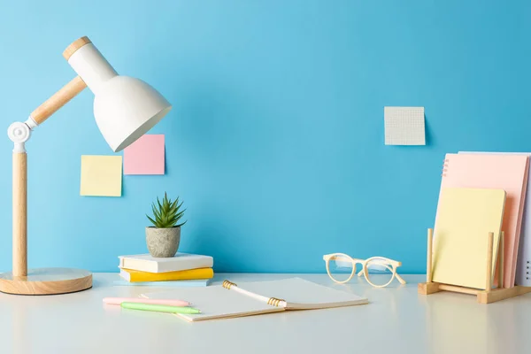 Academic essentials, such as books holder, copybook, glasses, and lamp neatly arranged on desk, captured in side view photo against blue wall with sticky notes. Perfect for text or promotional content
