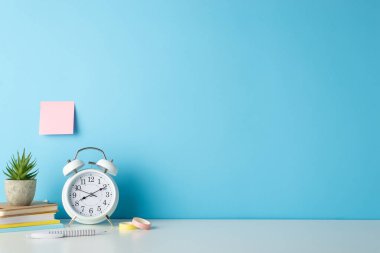 Beyaz bir masanın, çalar saatin, not defterlerinin ve kırtasiyenin yan açılı fotoğrafı vasıtasıyla öğrencinin çalışma alanındaki verimliliği mavi izole bir arkaplanda, metin veya reklamlar için idealdir